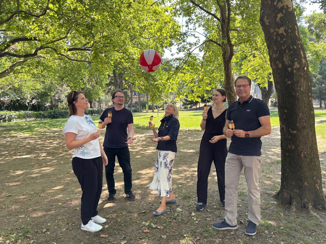 Fünf unserer Mitarbeiter:innen stehen mit einem Eis in der Hand im Park und werfen einen Wasserball in die Luft.